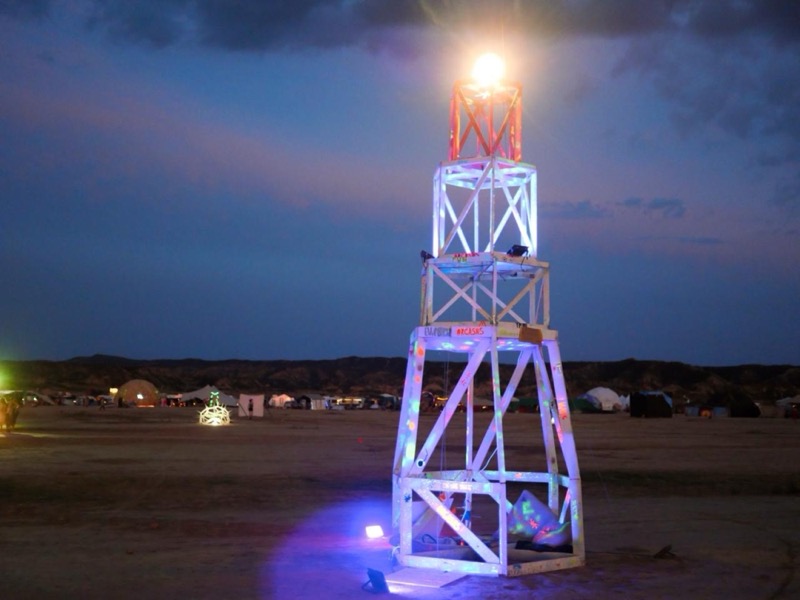 The lighthouse at dusk with its beacon lit, the camp and hills behind it
